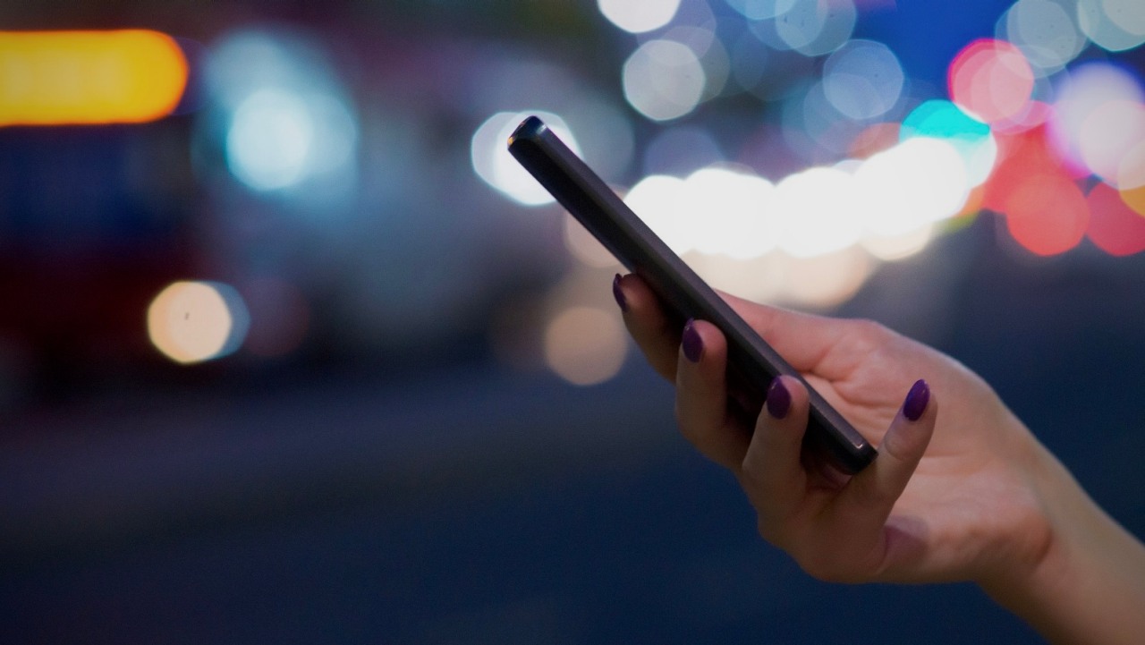 ORLEN innowacje. Kobieta trzyma w dłoni telefon komórkowy, w tle nadjeżdżający autobus.